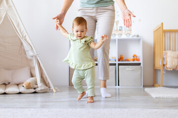 Baby girl learning to walk with mothers support at home. Lifestyle moment of childhood growth,...