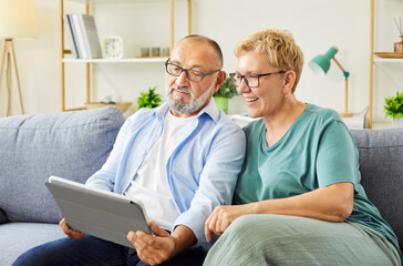 Elderly couple relaxed at home on a cozy sofa are using a tablet, browsing internet, smiling and...