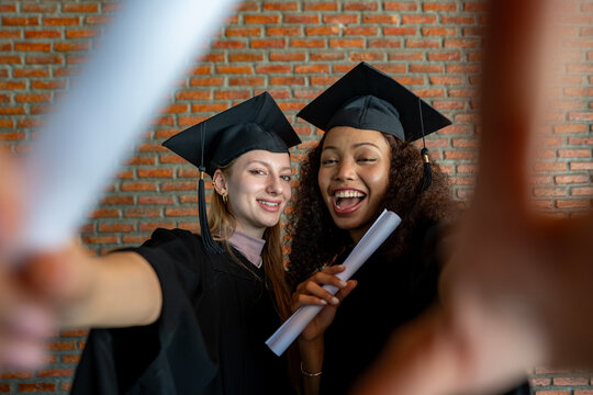 Portrait of college black and caucasian graduated students in graduation gown holding diploma taking selfie and smiling together for university education and success