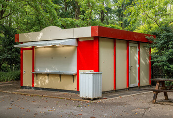 Closed outdoor food kiosk with red and beige walls, located in a green park surrounded by trees. The stand has a shuttered serving window.