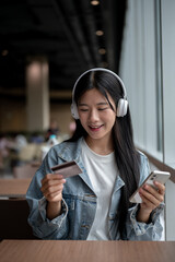 Young asian woman holding phone and looking at a credit card while sitting at table in cafe or...