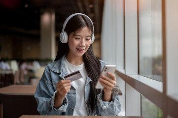 Young asian woman holding credit card and looking at phone while sitting at table in cafe or lounge.