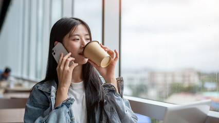 Young asian woman drinking coffee while holding phone talking aside laptop on table in the cafe.