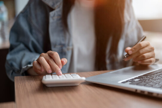 Close up of a woman's hands holding pen and using calculator aside laptop on wooden table in cafe.