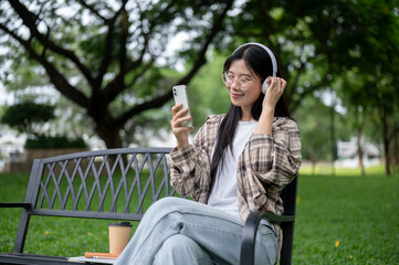 Young asian glasses woman looking at phone listening to music while sits on bench in park or garden.