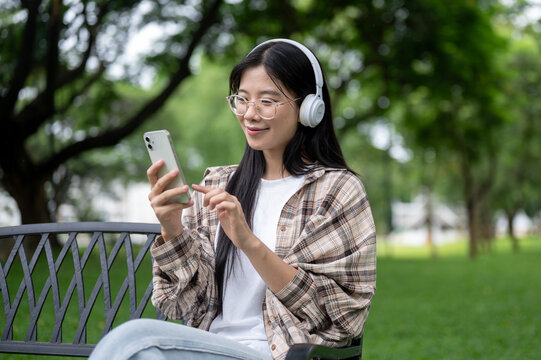 Young asian glasses woman holding phone listening to music in headphones while sits on bench in park