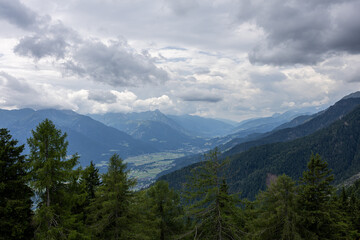 Wide view of alpine valley with mountains, forest slopes, and cloudy dramatic sky above