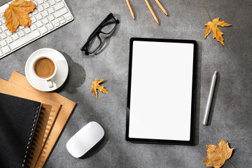 A cozy autumn workspace with mockup tablet with a blank screen, keyboard, mouse, coffee cup, notebook, pencils, glasses and fallen leaves on a gray stone desk. Top view. Copy space.