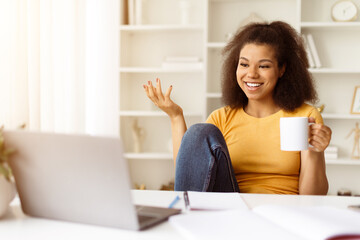 Smiling African American woman drinking coffee during video chat at laptop. Remote work, freelance lifestyle, online learning and modern home communication concept