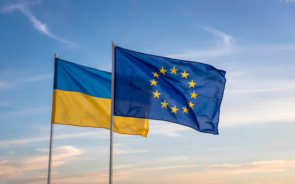 A photograph of two flags waving gently in a clear blue sky, the flags representing Ukraine and the European Union. The Ukrainian flag features horizontal bands of blue and yellow, while the European 