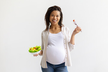 Smiling pregnant African American woman holding bowl of fresh salad and fork, standing against...