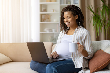 Pregnant African American woman holding white mug and working on laptop at home, smiling and relaxed, enjoying coffee or tea while sitting comfortably during pregnancy