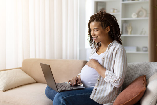 Smiling pregnant Black woman sitting on sofa at home with laptop, hand on belly, working online or browsing internet, enjoying lifestyle balance during pregnancy