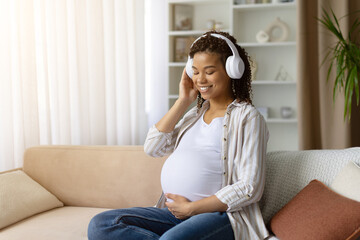 Smiling pregnant African American woman listening to music with headphones at home, relaxing on sofa with hand on belly, enjoying prenatal bonding lifestyle