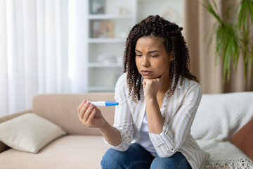 Concerned African American woman sitting on sofa at home, holding pregnancy test and looking at result with serious expression, uncertain about family planning future
