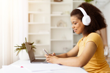 Focused African American woman with headphones typing on smartphone while studying at laptop. Online learning, remote freelance job or digital communication
