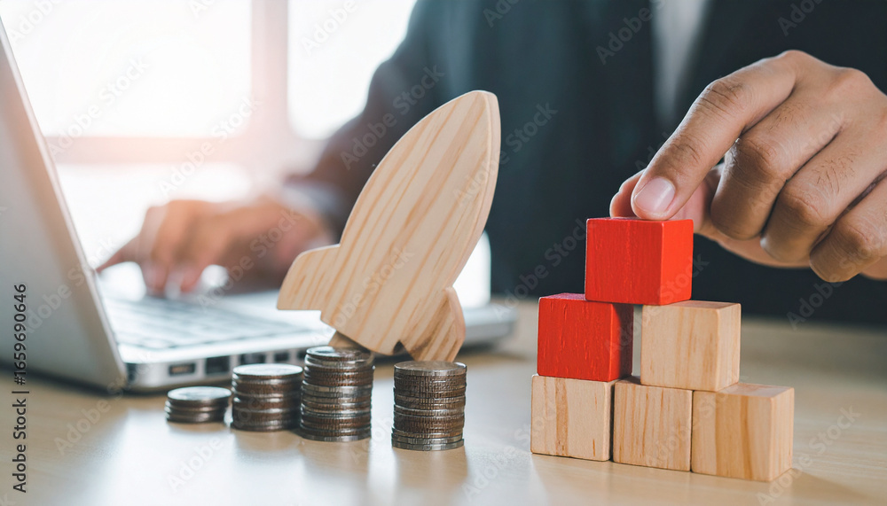 Poster a person building a tower with wooden blocks while stacks of coins and a rocket ship are in the fram