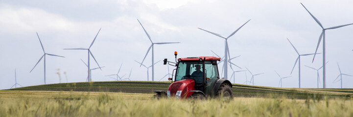 Red tractor navigates through whispering wheat fields beneath sentinels of wind power, symbolizing Earth Day's renewable promise