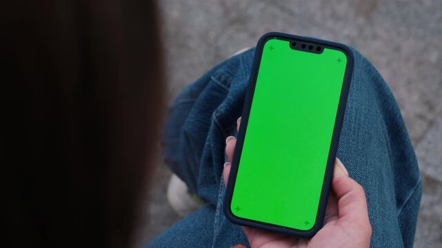 Woman holding smartphone with green screen mockup outdoor, sitting on the bench at the street using phone for chatting with friends, surfing social media, watching reels, video, reading book during re