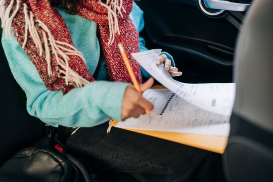 A person in a car is focused on filling out documents with a pencil. They are wearing a red scarf and a blue sweater, working on papers in a yellow folder.
