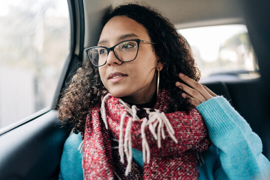 A young woman of color with curly hair and glasses sits in a car's back seat. She gazes thoughtfully out the window, her hand gently touching her hair. She wears a cozy red scarf and a blue sweater.