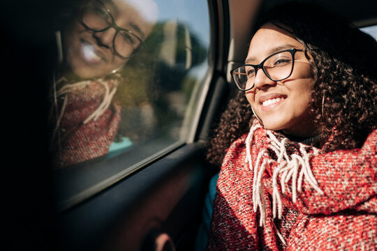 A smiling young woman with curly hair and glasses sits in a car, looking out the window. Her reflection is visible in the glass as sunlight illuminates her face.