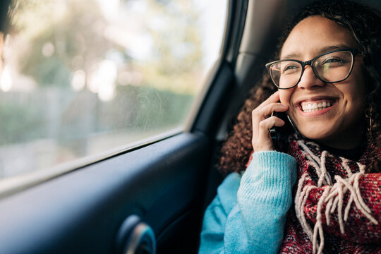 A happy young woman with curly hair and glasses is talking on her smartphone inside a car. She is smiling brightly while on the call.