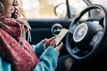 A woman with curly hair and glasses is seated inside a car, actively using her smartphone. She wears a warm red scarf and a blue sweater, focused on the device.
