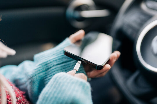 A person wearing a light blue sweater holds a modern smartphone. They are actively plugging a white charging cable into the device while inside a car.