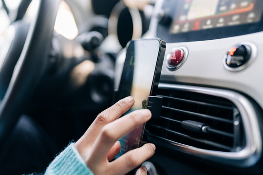 A person's hand adjusts a smartphone mounted on a car's air vent. The phone displays a screen, possibly for navigation or an application. The car interior is visible.