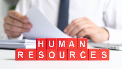 Obraz premium A close-up shot of the phrase HUMAN RESOURCES spelled out in red blocks, with a professional person in the background reading documents