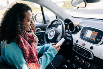 A woman with curly hair and glasses sits inside a car, focused on her smartphone. Her phone is connected to the vehicle, and the car's navigation system is active on the dashboard screen.