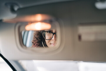 A woman with curly hair and glasses is reflected in a car's rearview mirror. Her eyes are visible through the glasses as she looks directly into the mirror.