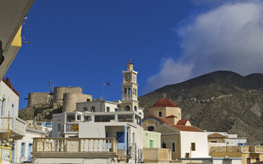 Greek village of Olympos, Karpathos island with whitewashed buildings, and a central clock tower. windmill stands in the background, with the Greek flag 