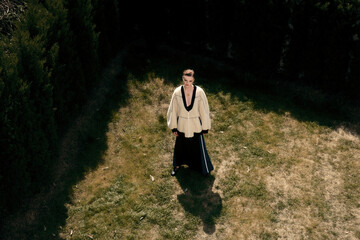 A 25-year-old woman in a stylish outfit standing on grass surrounded by greenery.