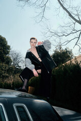Young white female model in oversized jacket poses on car roof.