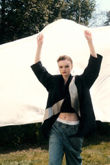 Young woman posing with raised arms in stylish outerwear against a draped fabric background.
