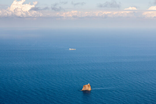 Aerial view of a blue sea with a small rocky island in the foreground and a cargo ship in the distance under a cloudy sky. Stromboli Island, Sicily, Italy
