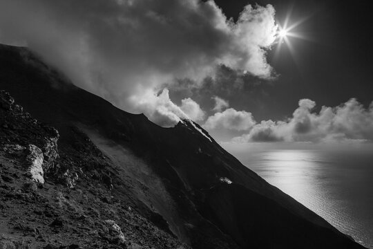 Black and white view of a volcano slope meeting the sea, with clouds and sun above. Stromboli Island, Sicily, Italy