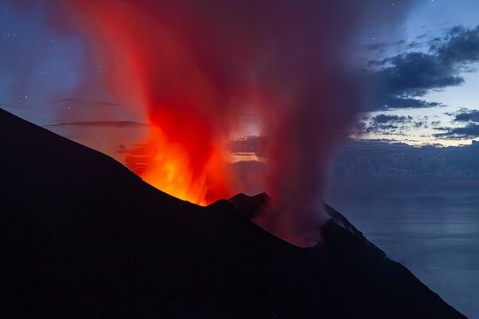 Night view of a volcano erupting, with bright orange lava and smoke against a dark sky and ocean. Stromboli Island, Sicily, Italy