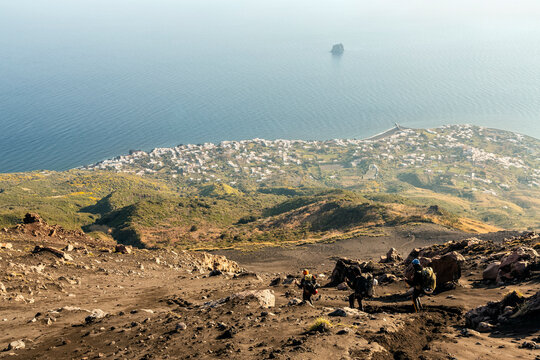 Three hikers descend a rocky, sandy slope, with a coastal town and the sea visible in the background. Stromboli Island, Sicily, Italy