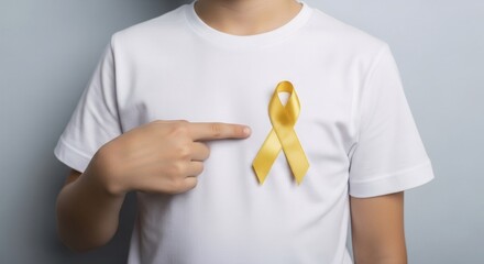 Caucasian boy stands with a yellow ribbon on a white t-shirt, pointing at it. Banner template for childhood cancer awareness month concept and support for disease patients. 