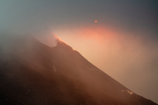 Volcano with smoke rising from the crater, a hazy sky, and the sun partially visible. The sea is on the right side of the image. Stromboli Island, Sicily, Italy