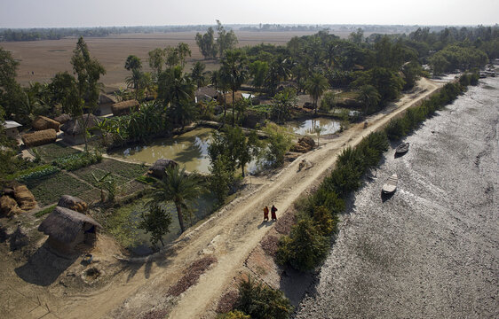 Aerial view of a sandy path winding through a village with ponds and thatched roofs, next to a muddy riverbank, Sundarban, West Bengal, India.