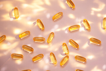 Yellow fish oil capsules on light pink background with sun glare and shadows.