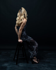 Young blond woman wearing elegant dress sitting on wooden chair in studio with dark background and...