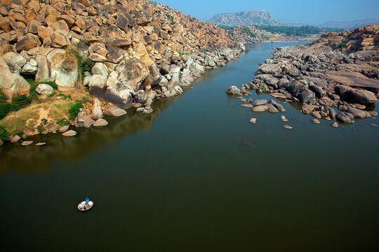 Aerial view of the Tungabhadra River winding through ancient granite boulders, a coracle drifts on the still, reflective water, Hampi, Karnataka, India.