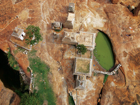 Aerial view of ancient structures nestled amidst the rugged, sun-baked landscape and an emerald pool, Badami, Karnataka, India.
