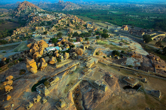 Aerial view of ancient ruins nestled among golden boulders and rugged hills under a soft sky, Hampi, Karnataka, India.