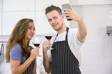 Happy couple taking selfie in modern kitchen. Man holding up smartphone to take picture while both holding cocktail glasses with red drink at home
